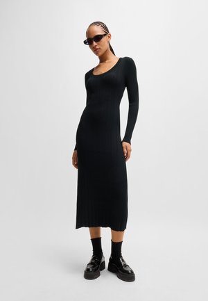Black ribbed long-sleeve dress with a scoop neckline, fitted silhouette, and a knee-length hem. Model wears chunky black platform shoes and sunglasses.