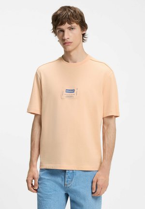 Młody mężczyzna o jasnobrązowych włosach, ubrany w brzoskwiniowy T-shirt z krótkim rękawem z niebieską naszywką z logo „HUGO” na środku klatki piersiowej oraz jasnoniebieskie jeansy.