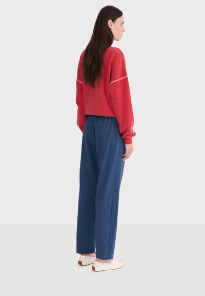 Kobieta z długimi ciemnymi włosami, ubrana w czerwony krótki sweter, luźne niebieskie spodnie i białe buty, stojąca bokiem na jednolitym tle.