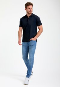 Mornarska polo majica s ozko prileganjem, kratkimi rokavi in ovratnikom, v kombinaciji s svetlo modrimi ozkimi jeans hlačami in belimi športnimi čevlji.
