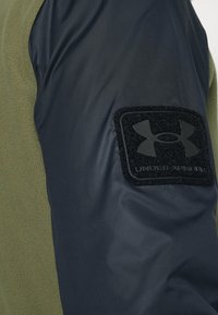 Fleecová bunda s olivově zeleným tělem a tmavě modrými rukávy. Na horní �části rukávu má černou nášivku s vylisovaným logem.