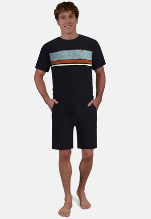 Lächelnder Mann steht barfuß, trägt ein schwarzes T-Shirt mit horizontalen blauen, orangefarbenen und weißen Streifen sowie schwarze Shorts mit Taschen.