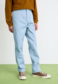 Lyseblå bukser med rett snitt, med en glatt tekstur, sammen med brune sneakers og en brun langermet topp. Grønn gulv bakgrunn.