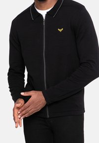 Homme portant un pull zippé noir avec un logo jaune brodé sur la poitrine, mains jointes devant, sur un fond blanc.