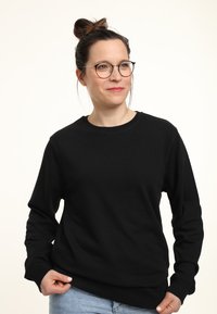 Jonge vrouw met een bril en haar in een knot, gekleed in een zwarte lange mouwen shirt en lichtblauwe jeans, die iets opzij kijkt.