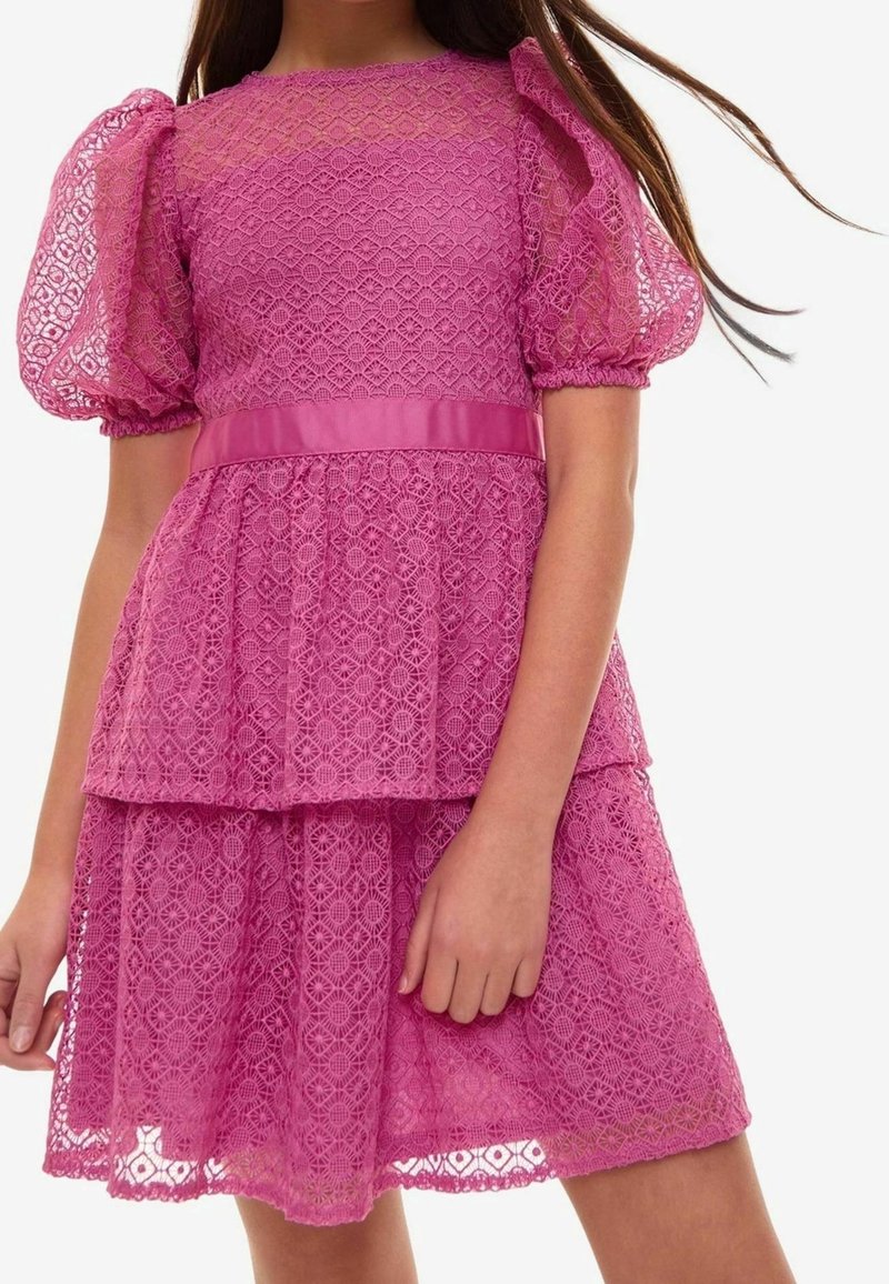Fille portant une robe en dentelle rose avec des manches bouffantes, une taille ajustée et une jupe à volants, se tenant devant un fond blanc uni.