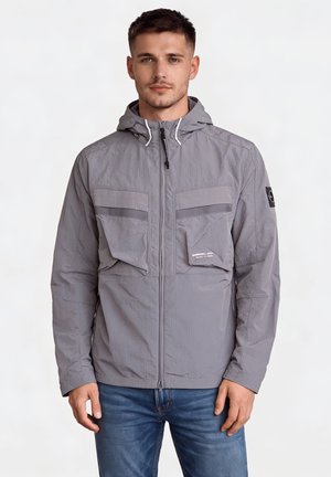 Jeune homme portant une veste à capuche grise avec zip et grandes poches avant, associée à un jean bleu, sur fond blanc.