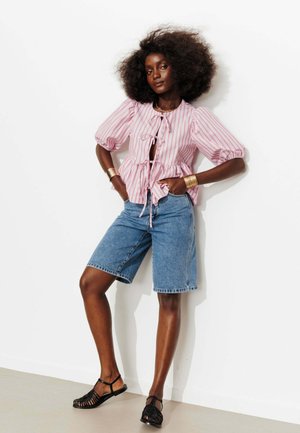 Femme aux grands cheveux bouclés portant un chemisier rayé rose et blanc, un short en jean jusqu’aux genoux, des sandales noires et des bracelets en or, debout contre un mur blanc.