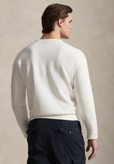 Moški, videti od zadaj, ki nosi belo srajco z dolgimi rokavi in temno mornarsko modre cargo hlače na nevtralnem ozadju.