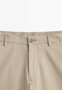 Beżowe spodnie z zapięciem na guzik i zamek błyskawiczny, z szlufkami na pasek i przeszyciami na jednolitym białym tle.