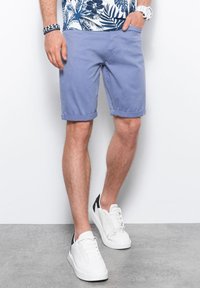 Pantaloni corti di cotone azzurri con risvolti, due tasche laterali, indossati con sneakers bianche con dettagli neri e una camicia a fantasia.
