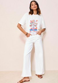 Weißes T-Shirt mit einem bunten Grafikdesign eines Blumenvasen, entspannter Schnitt, kombiniert mit weit geschnittenen weißen Hosen und braunen Riemchensandalen.