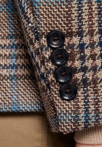 Brązowa marynarka w kratę houndstooth. Posiada cztery czarne guziki oraz teksturowany materiał z niebieskimi i kremowymi akcentami.