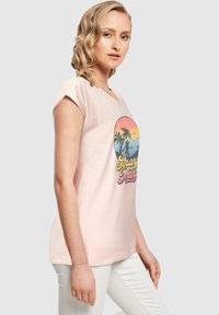 Lys rosa bomull t-skjorte med korte ermer, med et grafisk design av palmer og solnedgang. Tekst "Stranger Things" i gule og røde detaljer.