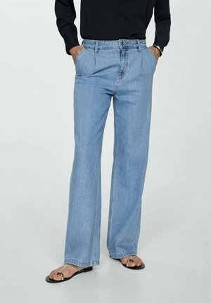 Personne portant un jean large bleu clair, un haut noir à manches longues et des sandales noires avec des anneaux dorés, mains dans les poches, debout sur un sol gris.