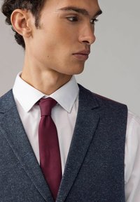 Navy teksturoitu liivi slim fit -mallilla, valkoisen paidan ja burgundin värisen solmion päällä. Huomattavina yksityiskohtina erottuva ompelu ja läpät.