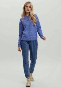 Paarse gebreide trui met een ronde hals, gedragen met blauwe taps toelopende jeans en beige enkellaarzen tegen een neutrale achtergrond.