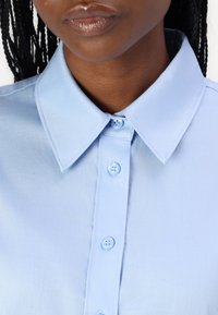 Camicia azzurra chiara con colletto in tessuto liscio, caratterizzata da una chiusura a bottoni con due bottoni visibili. Design semplice, senza motivi o accenti.