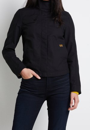 Vrouw draagt een zwart jack met rits en een klein borstzakje, samen met donkere spijkerbroek, hand in het voorzakje, tegen een effen witte achtergrond.