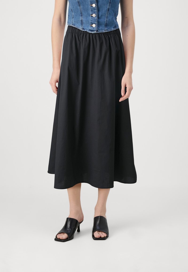 Femme portant une jupe midi noire avec une taille élastique, associée à des mules noires à talons ouverts et un top en denim sans manches.