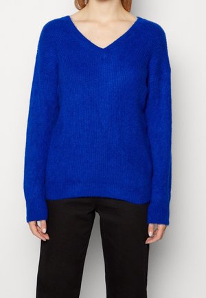 Pull V à col en V bleu vif en matière douce et texturée, avec des manches longues et une coupe décontractée, associé à un pantalon noir.