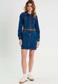 Robe en denim avec un devant boutonné, des manches longues et une taille ceinturée. Bleu foncé avec des coutures orange contrastantes. Associée à des baskets blanches.