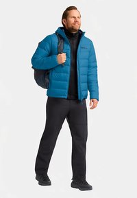 Tiefblaue Steppjacke mit Reißverschluss vorne und Kapuze, über einem schwarzen Langarmshirt. Schwarze Hose und Schuhe, dazu ein grauer Rucksack. Glatte Textur.