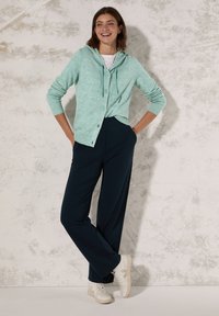 Ljus teal kofta i stickad design med huva, knappar framtill och ribbade ärmslut. Matchad med marinblå vida byxor och vita sneakers.