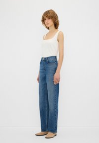 Jeans in denim blu a vita alta con una vestibilità dritta, caratterizzati da un girovita con chiusura a bottone frontale e dettagli di cucitura sottili, abbinati a una canotta bianca.