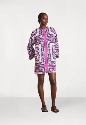 Femme portant une robe longue à manches longues, mi-longue, aux motifs roses et violets, avec des sandales marron, debout devant un fond blanc uni.