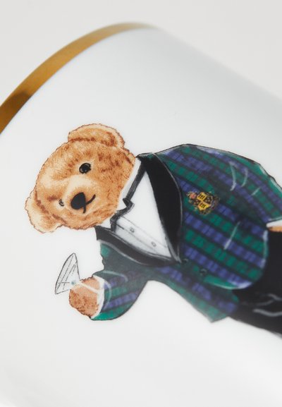 Mali medvjed u zeleno-plavom kariranom smokingu drži martini čašu na bijeloj pozadini s zlatnim rubom.