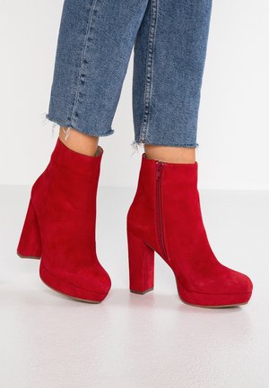 Personne portant un jean court bleu et des bottines à talons hauts en daim rouge vif avec des fermetures éclair latérales sur un sol lisse.