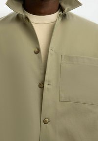 Gros plan d'une personne portant une chemise kaki à boutons sur un pull en tricot beige, mettant en valeur les boutons de la chemise et la poche poitrine.