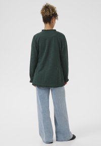 Zelený texturovaný top s dlouhými rukávy a volánkovým lemem, doplněný světle modrými širokými kalhotami. Jednoduchý design bez viditelných vzorů či kovových doplňků.