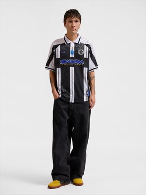 Jeune personne debout, les mains dans les poches, portant un maillot de sport rayé noir et blanc, un pantalon noir ample et des chaussures jaune-brun sur fond uni.