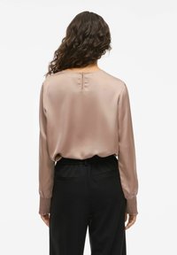 Žena s dlhými kučeravými vlasmi, oblečená v mauve saténovej blúzke so zhupovanými manžetami a čiernymi nohavicami s vysokým pásom, zobrazená zozadu.