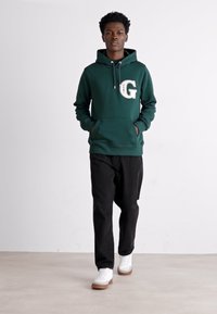 Felpa verde con cappuccio caratterizzata da un grande logo bianco "G", abbinata a pantaloni neri e sneakers bianche, su uno sfondo neutro.