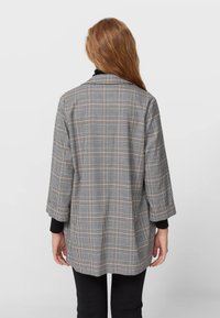 Grijze geruite blazer met driekwart mouwen, gemaakt van een gestructureerde stof. Heeft een losse pasvorm met een scherpe kraag en zwarte accenten.