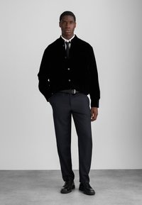 Black velvet jacket with a collar, front buttons, and pocket, paired with dark trousers and black lace-up shoes. Minimalist design and sleek texture.