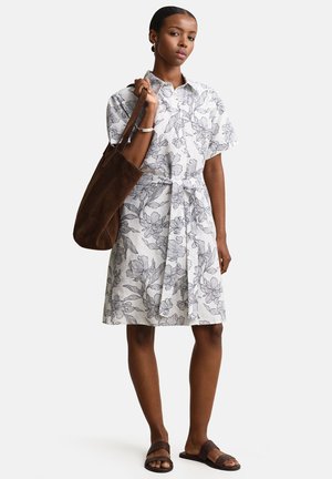 Femme portant une robe blanche à manches courtes à motifs floraux, tenant un grand sac à bandoulière marron et portant des sandales tissées foncées.