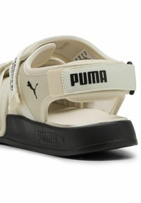 Beige sandaler med en stofrem og blød polstret sål. Har et sort logo på hælen og en struktureret sort gummiydersåle.