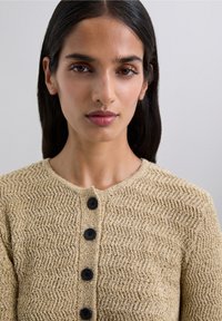 Young woman with long dark hair wears beige textured knit cardigan with five black buttons against plain background.