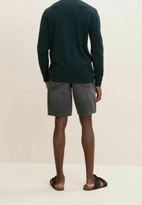 Donkergroene gebreide trui, grijze denim shorts, zwarte sandalen. Achteraanzicht toont eenvoudig ontwerp, geribbelde manchetten en detail van de achterkant van het juk.