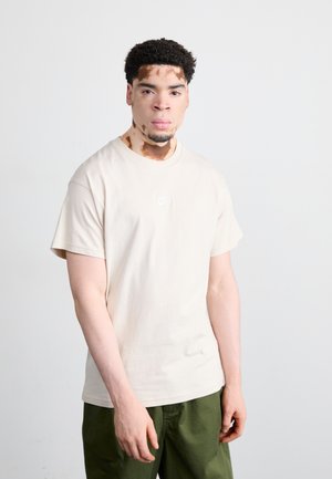 Beige cotton t-shirt with a crew neck and short sleeves, featuring a small white Nike logo on the chest. Worn with green pants.