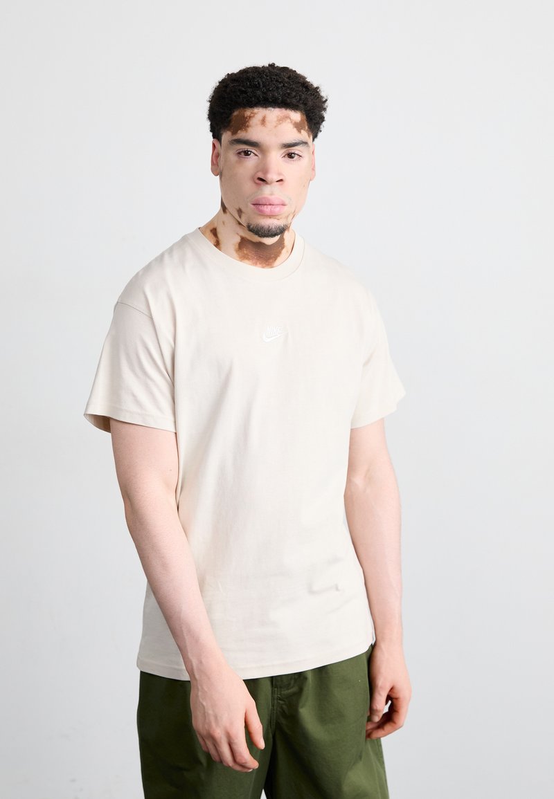 Μπεζ βαμβακερό t-shirt με στρογγυλή λαιμόκοψη και κοντά μανίκια, με μικρό λευκό λογότυπο Nike στο στήθος. Φορεμένο με πράσινα παντελόνια.