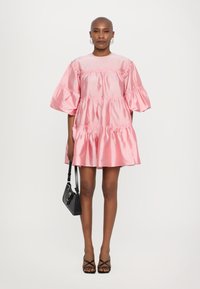 Ροζ σατέν φόρεμα με κοντά φουσκωτά μανίκια και σχεδίαση με σειρά ραβδώσεων. Το μοντέλο κρατά μια μαύρη τσάντα. Λείος υφασμάτινος χαρακτήρας, μήκος πάνω από το γόνατο.