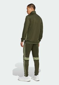 Olive green tracksuit made of smooth, stretchy fabric; features white stripes on sleeves and pants; zip collar; fitted design; paired with white sneakers.