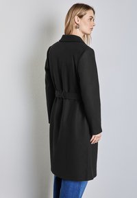 Czarny długi płaszcz z kołnierzem, przewiązany w talii, z długimi rękawami. Gładka tekstura i czyste linie, noszony na niebieskim dżinsie.