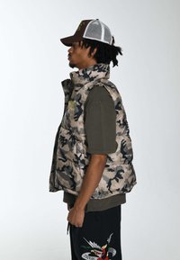 Kamuflażowa kamizelka puchowa z wysokim kołnierzem, wyściełanym wzorem i zapięciem na zamek, w połączeniu z jednolitą oliwkową koszulą T-shirt i ciemnymi spodniami z haftem.