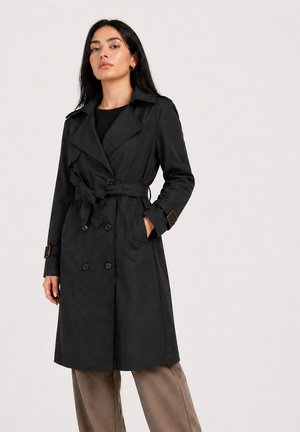 Kvinde stående iført en sort trenchcoat med bælte og knapdetaljer samt brune bukser mod en ensartet lys baggrund.